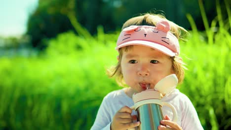 adorable american child girl drinking from kids' water bottle in a park against lush green bushes - slow motion, portrait