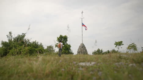 hiker with a backpack walking towards the rock structure with a slovenian flag and a cross at the top of a pole