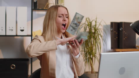 Cheerful-rich-business-woman-working-on-office-laptop-wasting-throwing-money-to-camera-profit-win