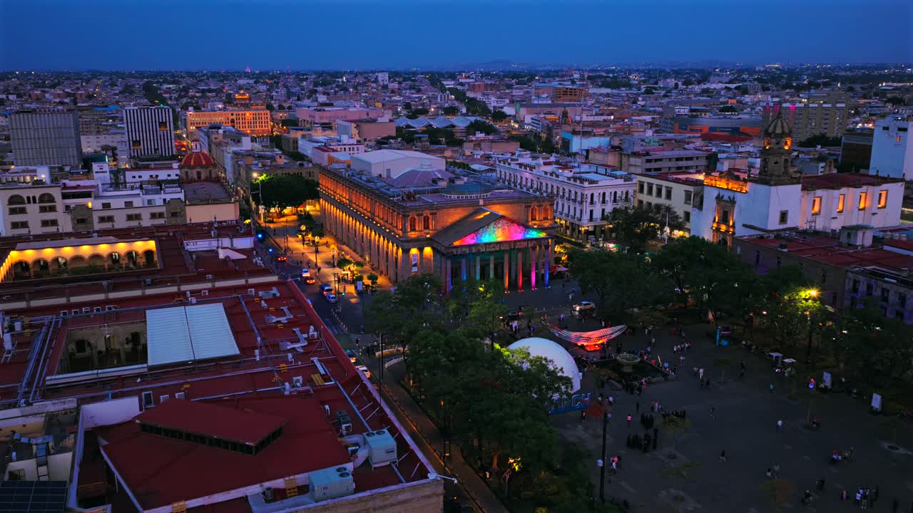 Drone captures vibrant evening aerial view of Guadalajara's Teatro Degollado, beautifully lit with colorful lights, surrounded by urban cityscape and lively plaza below
