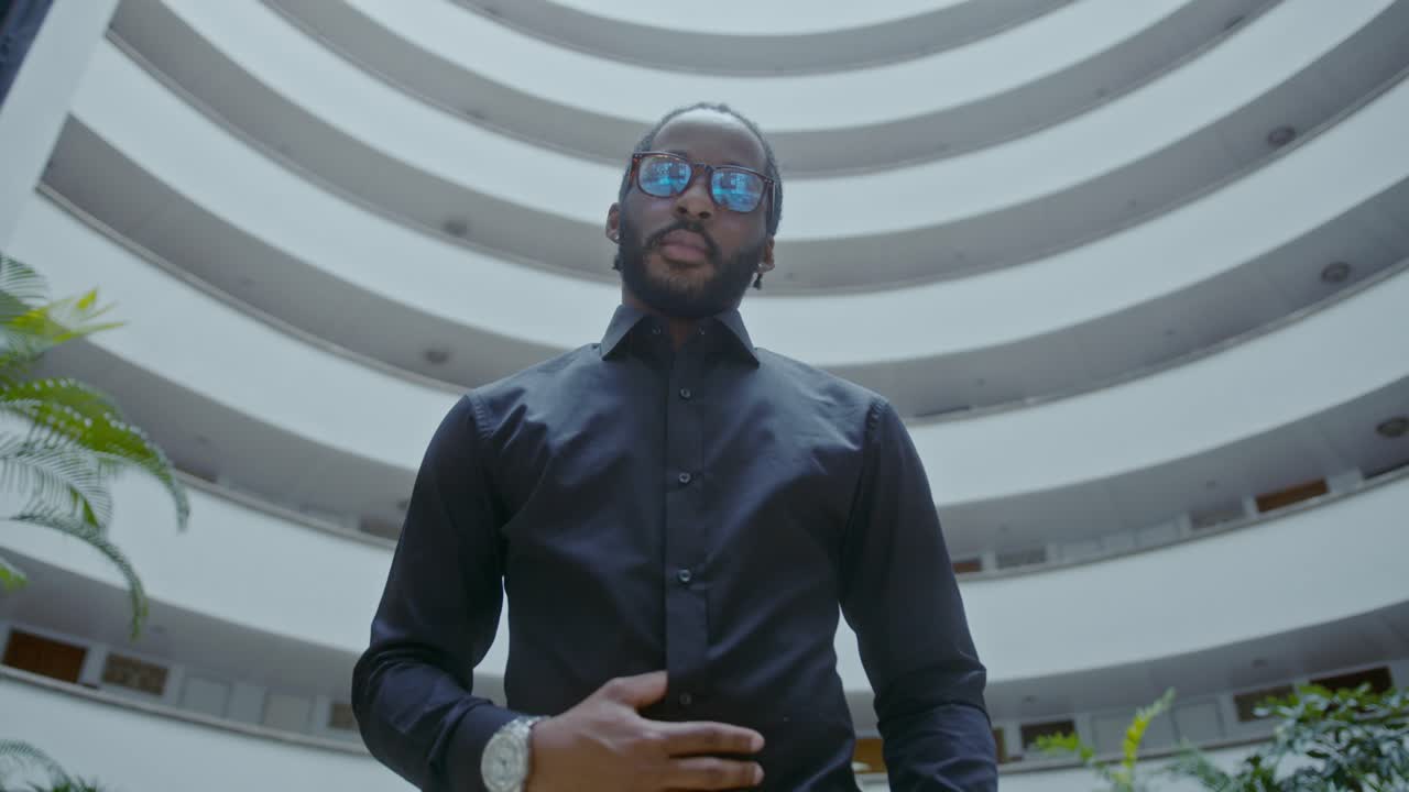 A man in a black shirt and eyeglasses stands in a modern building interior