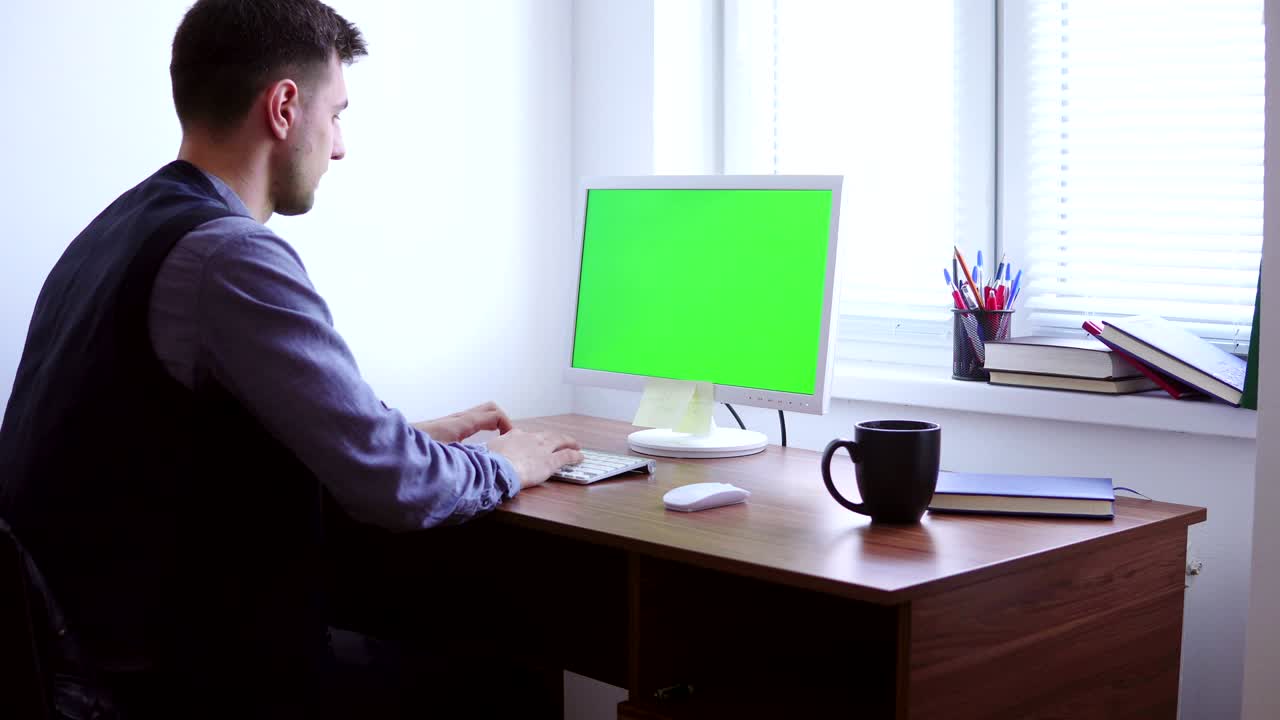 젊은이가 창 에 있는 테이블에서 녹색 화면 모형으로 컴퓨터에서 일하고 있다. 백인 노동자가 키보드에 타이핑하고 녹색 스크린을 보고 있다.