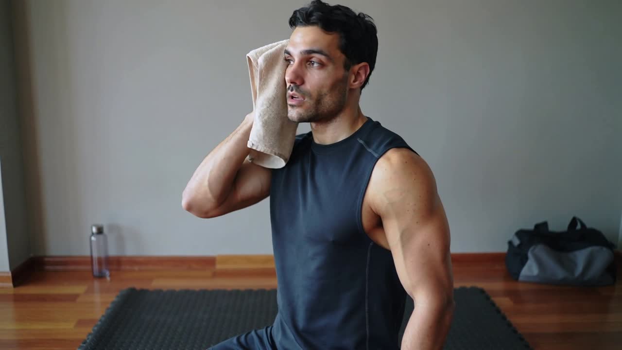 Pria bugar berbaju hitam tanpa lengan mengusap wajahnya dengan handuk setelah berolahraga di gym rumah.