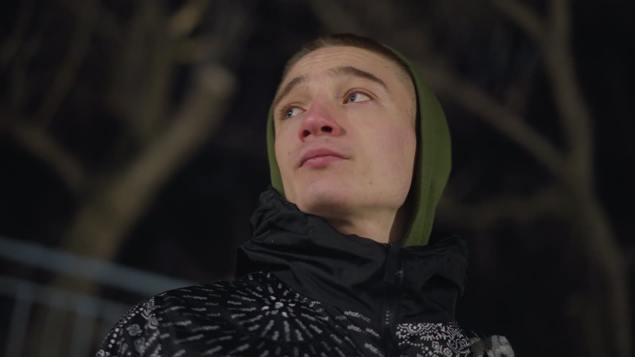 Side view of young boy in hoodie and patterned jacket gazing thoughtfully into distance during cold night, with leafless trees forming moody background