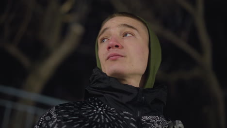 side view of young boy in hoodie and patterned jacket gazing thoughtfully into distance during cold night, with leafless trees forming moody background