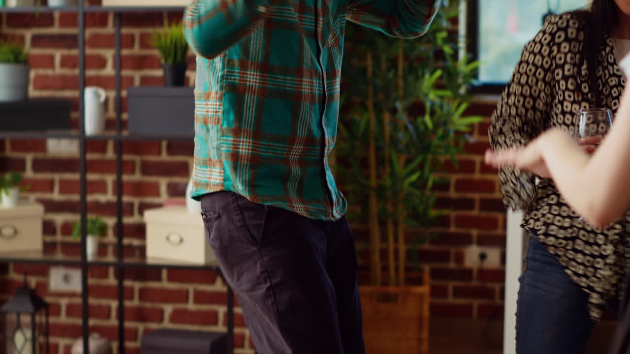 Guests at home party doing dance moves in brick wall living room, holding wine glasses in hand