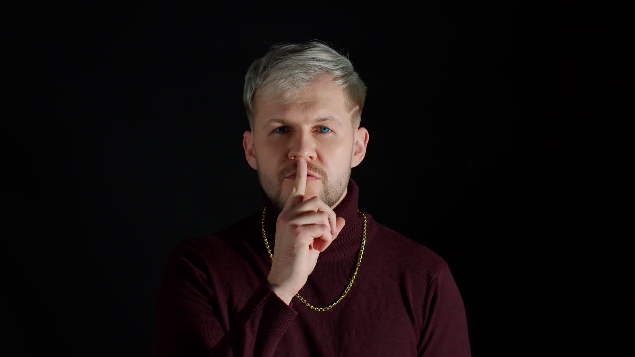 Blonďatý muž s modrýma očima přikládající si prst na rty