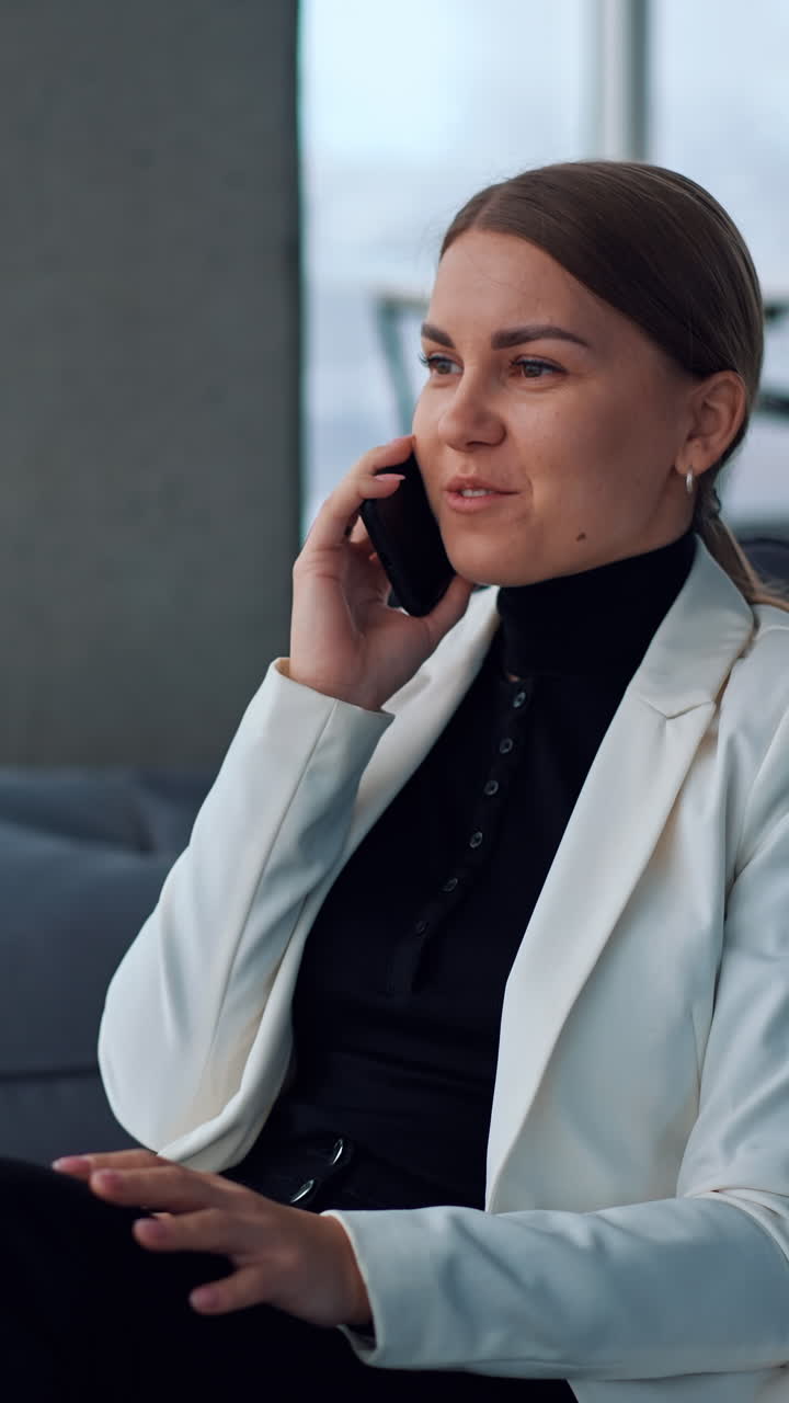 Woman having a break from work and speaking on the phone. Relaxed smiling lady sitting in bean bag chair and looking into window chatting on the smartphone. Vertical video
