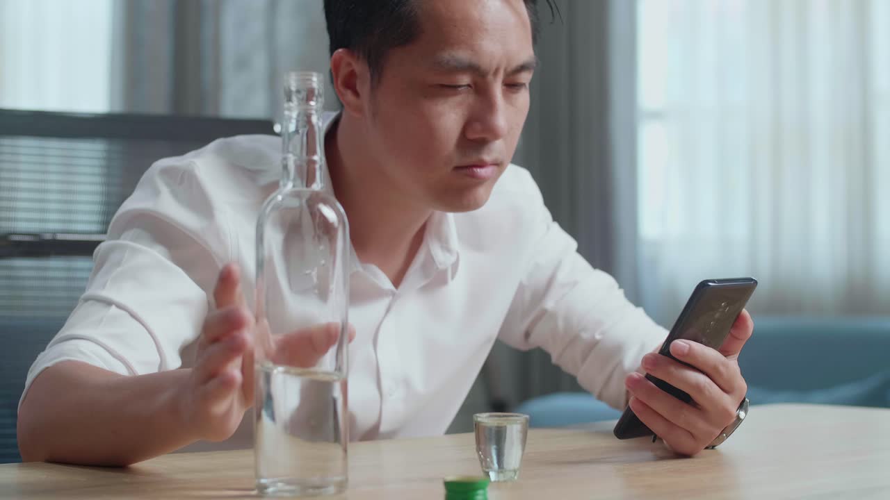 술에 취한 우울한 아시아 남자가 집에서 스마트폰을 사용하는 동안 술을 마시기 전에 글라스에 보드카를 부어 놓습니다.