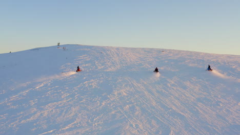vista aérea moto de nieve acelerando ladera nevada iluminada por el sol en el círculo polar ártico con un grupo de amigos