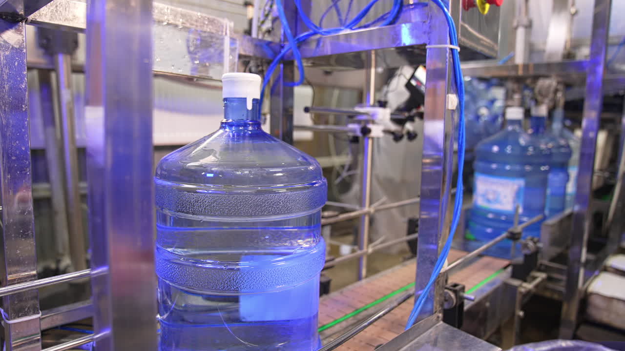 Big full bottles of clean water move by the conveyor belt. Machine is lifting a bottle and worker covers it in plastic bag.