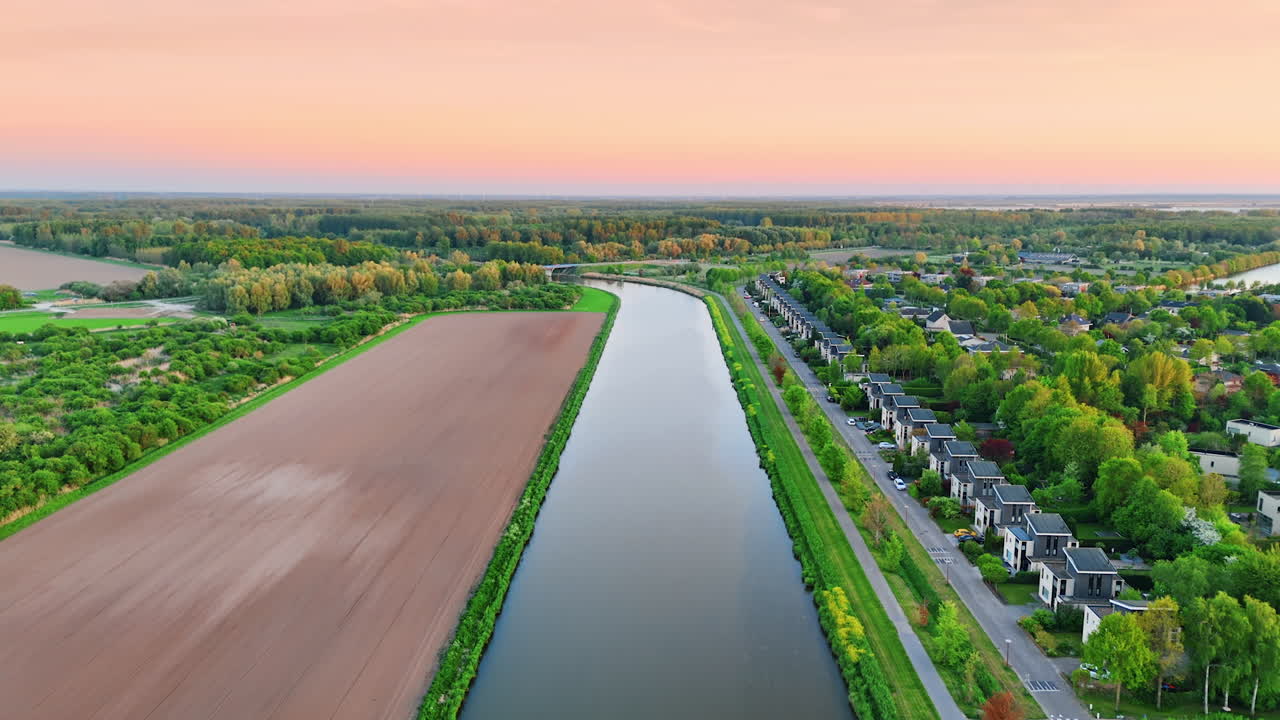Flying over the smooth surface of canal near the agricultural field. Revealing view on the cozy village and spectacular nature of the Netherlands.