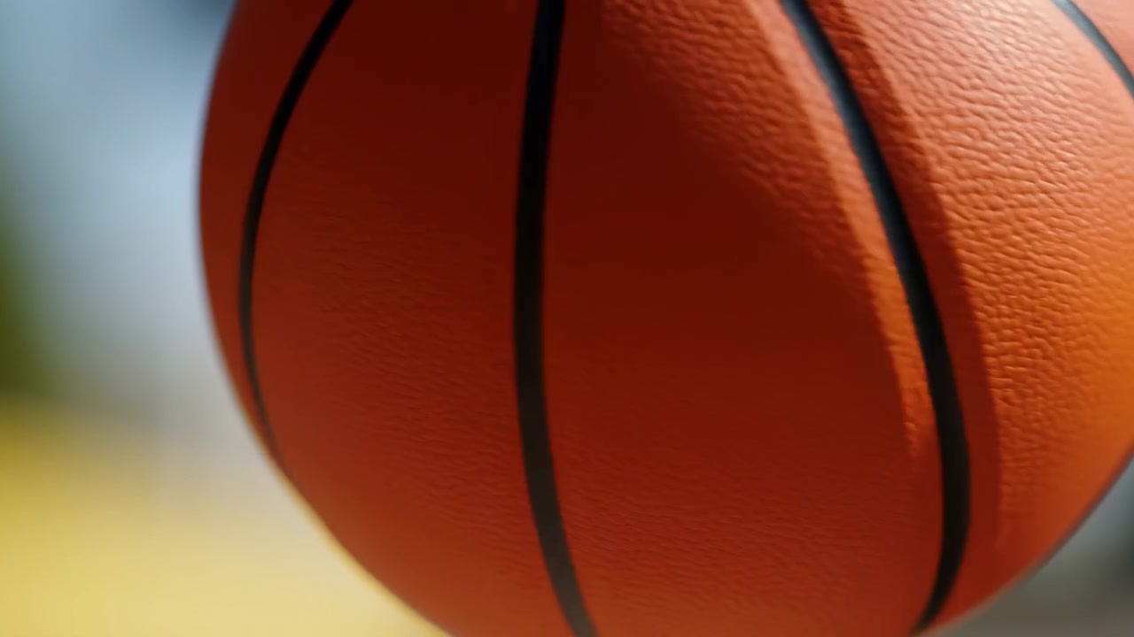 Close-up miring ketat bola basket oranye yang menunjukkan tekstur kulit berbutir kerikil yang detail dan beberapa jahitan hitam, dibingkai terpotong melintasi gambar dengan kedalaman bidang dangkal