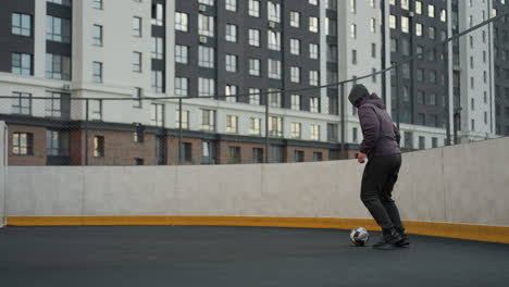 joven atleta controlando hábilmente la pelota de fútbol con los pies en la arena deportiva durante una sesión de entrenamiento, el fondo muestra edificios urbanos modernos, cercas de enlaces de cadena y iluminación vibrante