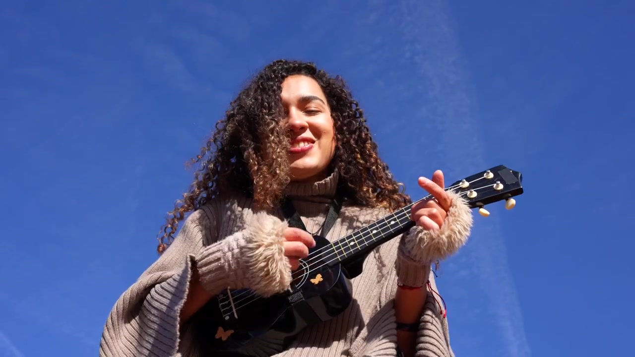 Young curly haired musician happily playing ukulele and singing outdoors against a clear blue sky