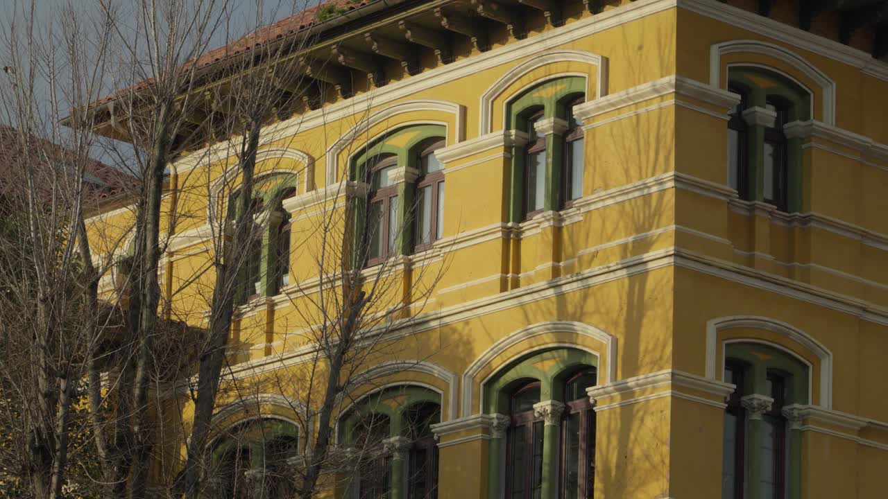 Bella architettura coloniale gialla con finestre ad arco decorate e rami spogli a Gijón, Spagna, durante il tramonto dell'ora d'oro. Facciata storica di un edificio europeo con elementi di design classici.