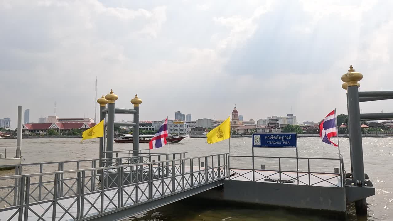 A tranquil scene at a Bangkok pier with flags waving and cityscape in the background under soft daylight