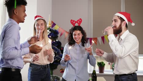 Group-of-happy-office-workers-holding-Bengal-lights-and-dancing-wearing-christmas-hats-and-deer-headband-and-celebrating