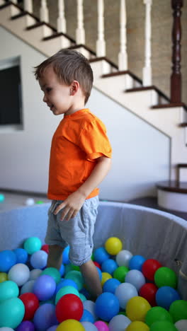 energetic baby boy tumbling in the colorful balls in the dry pool. happy kid stands up and tosses the balls. vertical video.