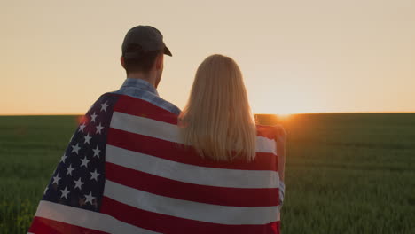 young couple with usa flag on their shoulders admiring the sunrise over the wheat field