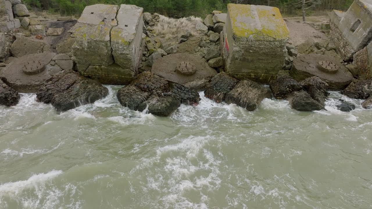 Aerial establishing view of abandoned seaside fortification buildings at Karosta Northern Forts, Baltic sea coast , large waves, overcast day, slow motion, drone shot moving backward