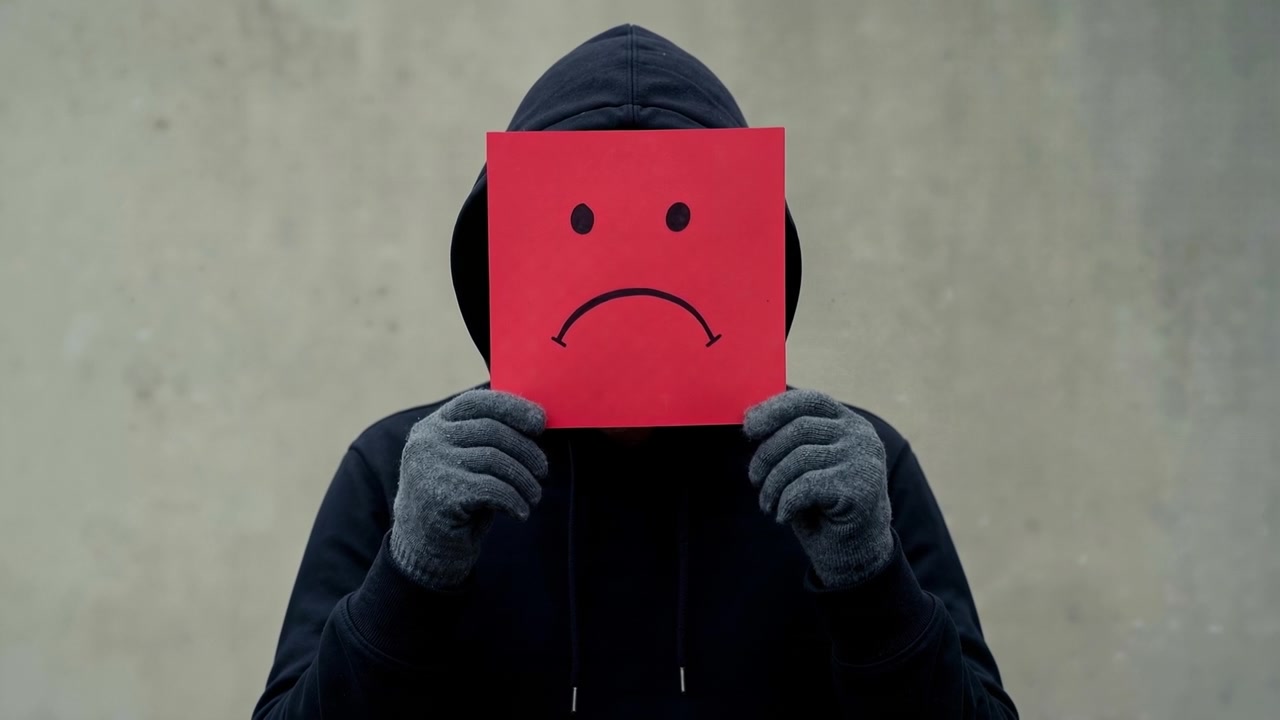 Person wearing a black hoodie holding a red sad face mask in front of their face