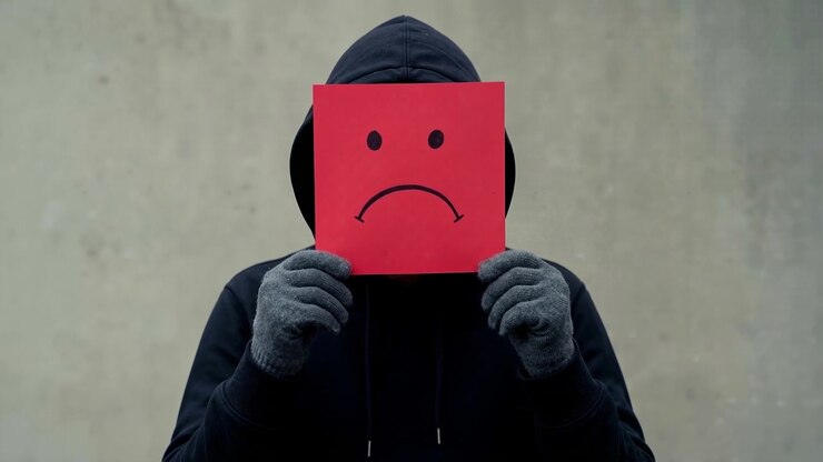 Seseorang mengenakan hoodie hitam memegang masker wajah sedih berwarna merah di depan wajahnya.