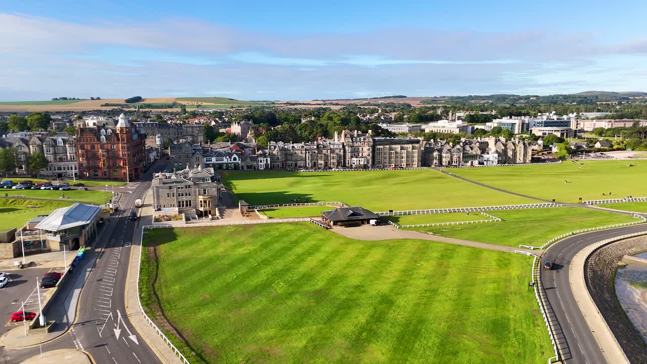 Drone camera glides above St Andrews, Scotland, revealing Victorian architecture, green golf course, sandy beach, and bright daylight with clear skies