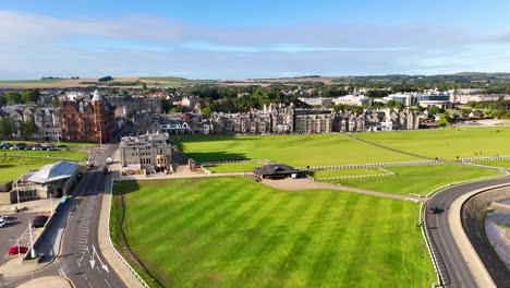 drone camera glides above st andrews, scotland, revealing victorian architecture, green golf course, sandy beach, and bright daylight with clear skies