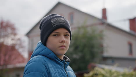 close-up of boy wearing black beanie and blue jacket standing outdoors, looking thoughtful and serious, with blurred background of red-roofed house and vibrant autumn foliage