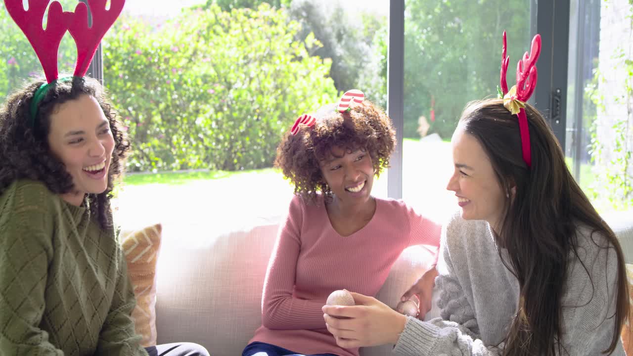 When left woman showing ornament, diverse women wearing antlers passing it pretending nose on couch