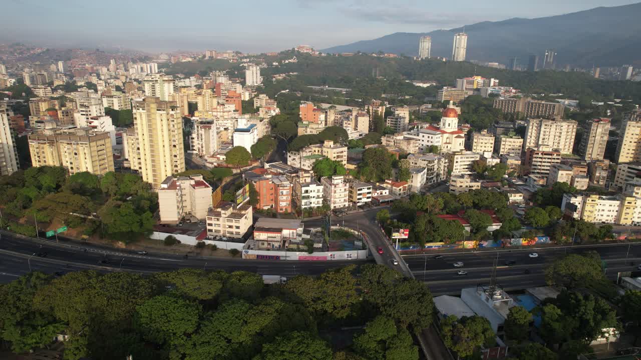Una città soleggiata con parchi verdi, edifici alti e montagne sullo sfondo, vista aerea.