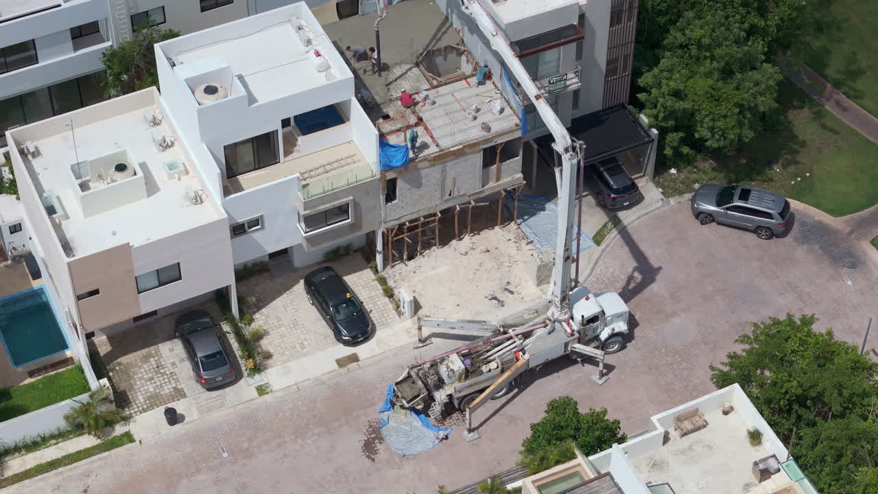 Panning tilt down drone shot of a concrete pump pouring cement to make the tile of a house with several workers spreading the material with shovels
