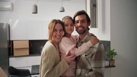 retrato de una familia feliz después de mudarse un hombre moreno con paja sostiene a su hija pequeña en ropa rosada en sus brazos y son abrazados por una chica morena en un suéter beige mientras están en un apartamento moderno después de moverse