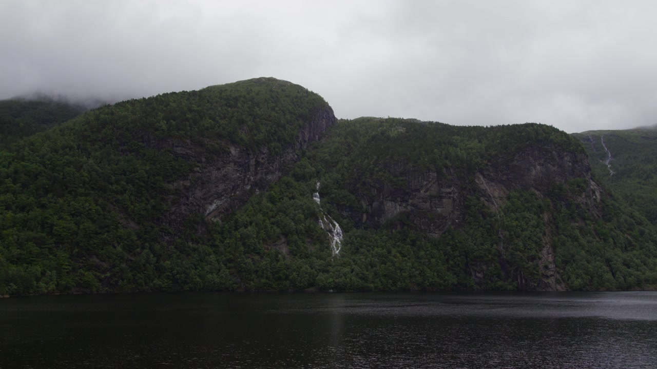 Una stretta cascata bianca scende da una ripida montagna verde in un fiordo immobile sotto cieli coperti, utile come inquadratura di apertura di una tranquilla natura nordica.