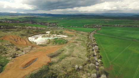 Toma-Aérea-De-La-Cantera-De-Caolín-Con-Un-Lago-Y-Campos-Verdes-Y-Montañas-En-El-Fondo,-Bajo-Nubes-De-Tormenta-Dramáticas