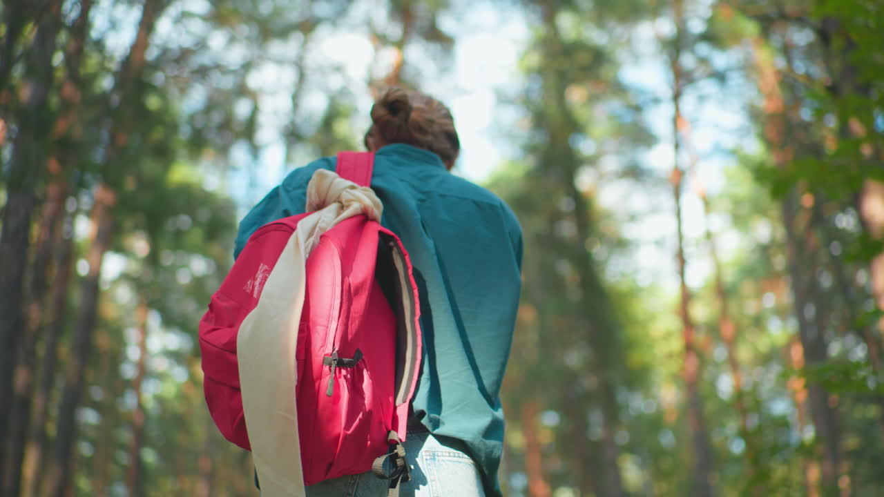Back view of woman walking through peaceful sunlit forest, red backpack draped over her shoulder, green shirt blending with nature as sunlight filters through the trees
