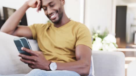 Feliz-Afroamericano-Relajándose-En-El-Sofá-Usando-Un-Teléfono-Inteligente-Y-Sonriendo-En-La-Sala-De-Estar,-En-Cámara-Lenta