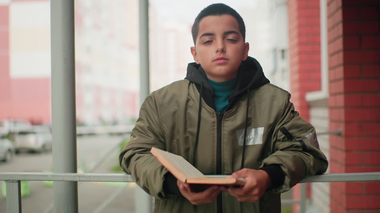 Little boy in green jacket holds open book while gazing thoughtfully, standing near brick wall with blurred city background and parked cars creating soft urban atmosphere