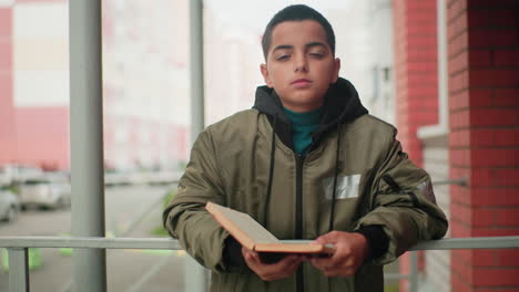 little boy in green jacket holds open book while gazing thoughtfully, standing near brick wall with blurred city background and parked cars creating soft urban atmosphere