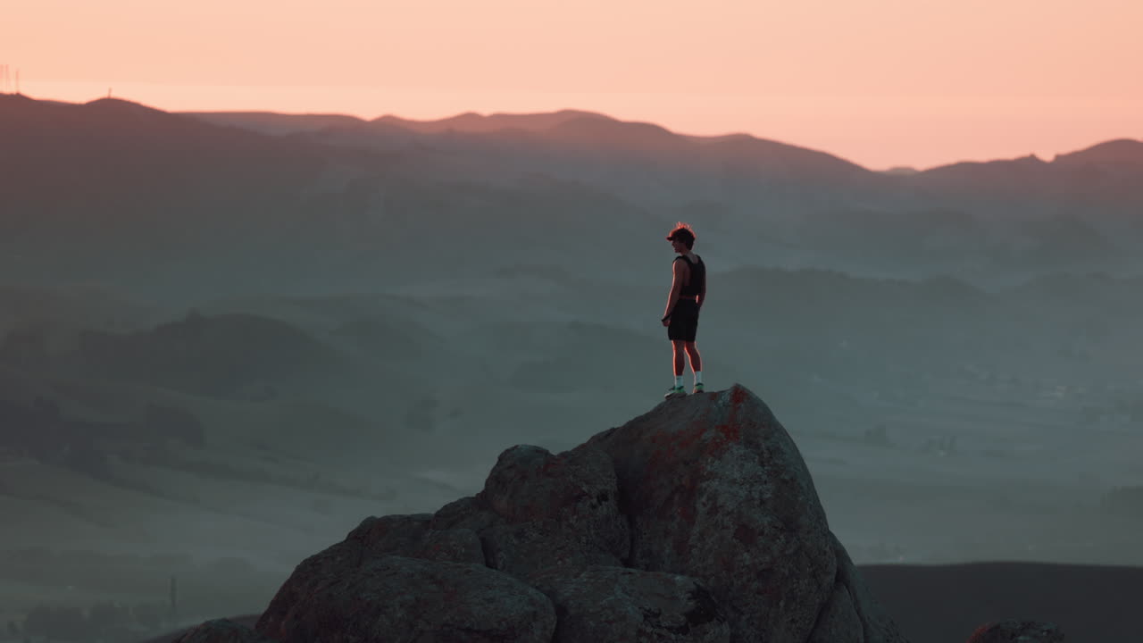 Teenager stands on cliff and looks out at vast rolling hills and mountains during sunset. Shot on Canon R5 at 4k 120fps with RF 70-200mm f2.8.