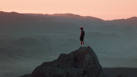teenager stands on cliff and looks out at vast rolling hills and mountains during sunset. shot on canon r5 at 4k 120fps with rf 70-200mm f2.8.