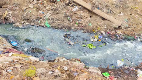 sewerage spews out of pipe surrounded by garbage and flows along a filthy mud bank past a scavenging dog into a river in bangladesh