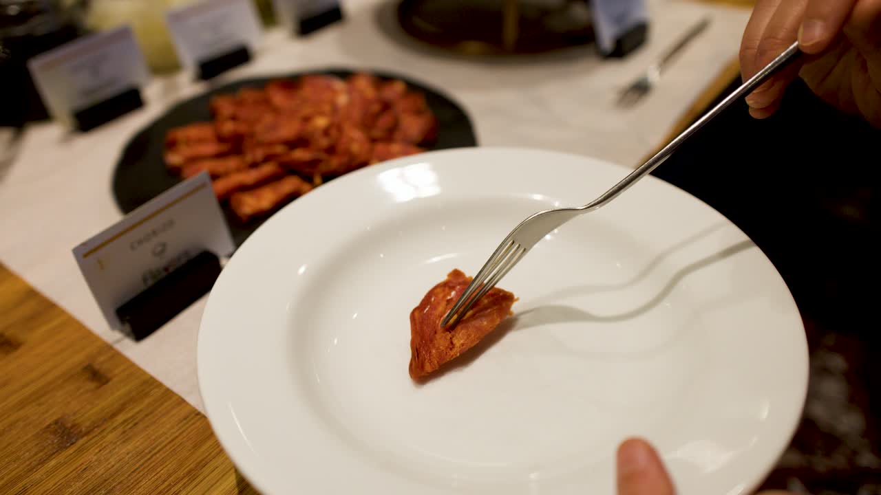 A hand uses a fork to place sliced chorizo onto a white buffet plate