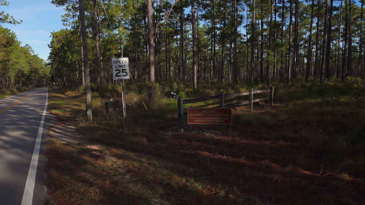 Drone trækker sig tilbage fra skovrydning til 25 mph fartbegrænsning og Blackwater River State Park-skilt, blandede træer og underskov i varmt eftermiddagslys