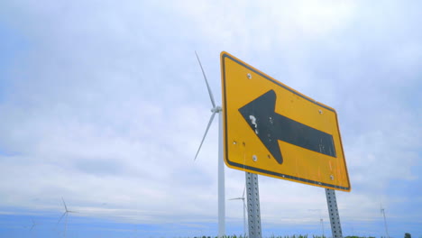 Road-sign-pointing-to-wing-turbines-field-against-clouds-sky