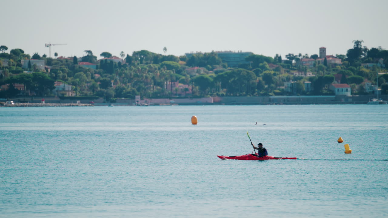 A man paddles a red kayak across the calm turquoise sea near a coastal town