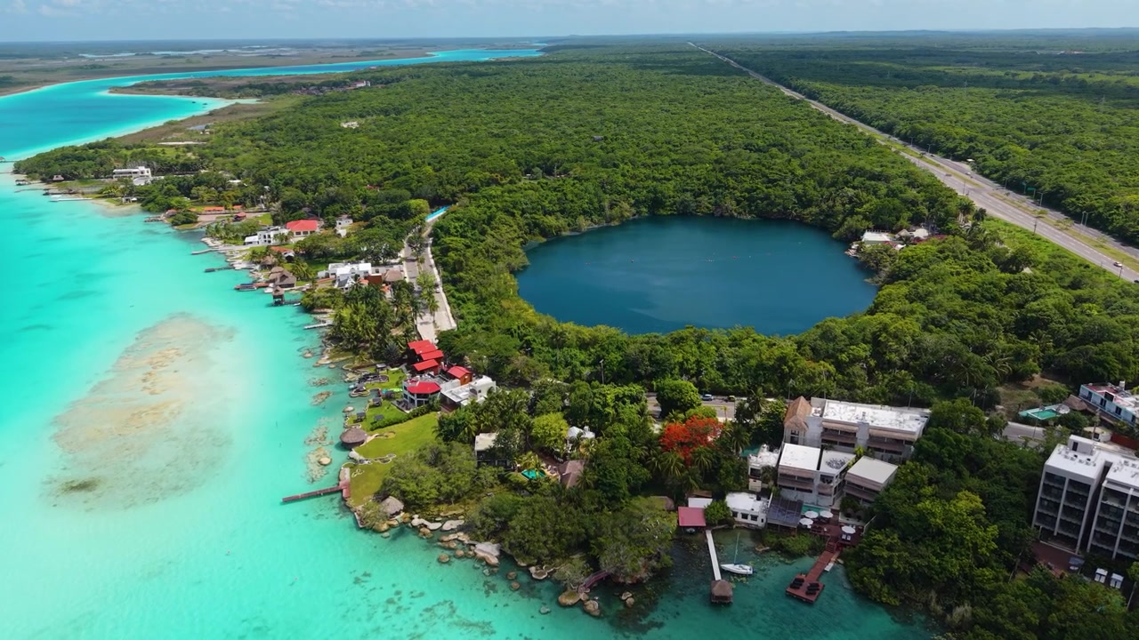 A deep blue sinkhole sits beside shallow aquamarine waters and lush green forest canopy.