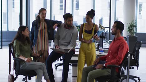 Diverse-group-of-work-colleagues-holding-laptop,-tablet-and-documents-talking-and-smiling-to-camera