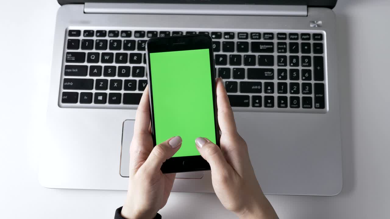 Female hands typing on a big black smartphone, Green screen, chromakey concept, laptop in the background. Top shot 60 fps